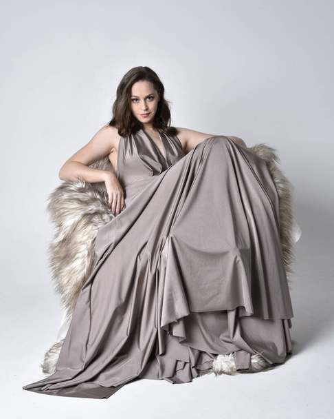 Retrato de uma menina bonita morena vestindo um longo vestido de noite de prata, comprimento total sentado pose na cadeira contra um fundo de estúdio
. - Foto, Imagem
