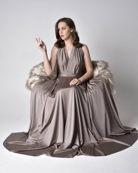 Portrait of a pretty brunette girl wearing a long silver evening gown, full length sitting pose on chair pose against a studio background. - Fotografie, Obrázek