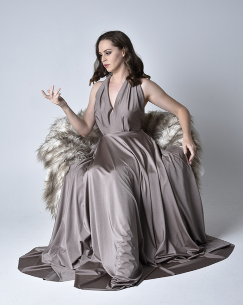 Porträt eines hübschen brünetten Mädchens, das ein langes silbernes Abendkleid trägt, in voller Länge sitzend auf einem Stuhl vor Studiohintergrund posiert. - Foto, Bild