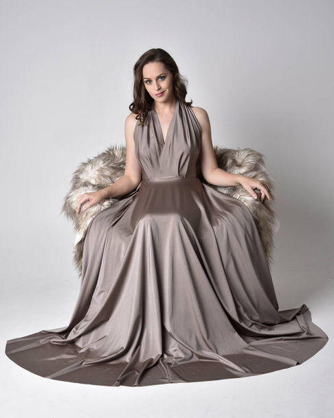 Porträt eines hübschen brünetten Mädchens, das ein langes silbernes Abendkleid trägt, in voller Länge sitzend auf einem Stuhl vor Studiohintergrund posiert. - Foto, Bild
