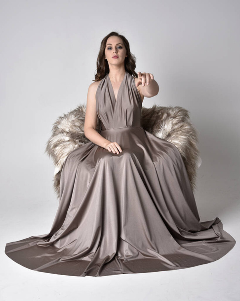 Portrait of a pretty brunette girl wearing a long silver evening gown, full length sitting pose on chair pose against a studio background. - Photo, Image