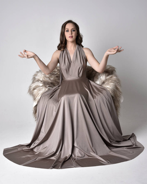 Portrait of a pretty brunette girl wearing a long silver evening gown, full length sitting pose on chair pose against a studio background. - Photo, Image