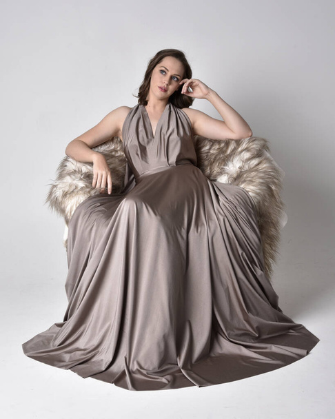 Portrait of a pretty brunette girl wearing a long silver evening gown, full length sitting pose on chair pose against a studio background. - Photo, image