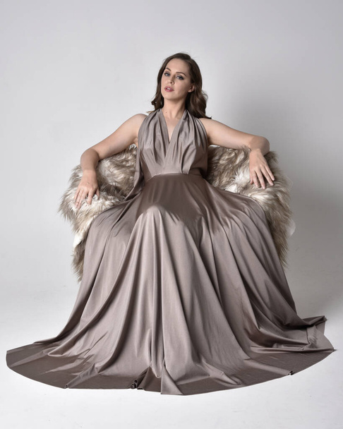 Portrait of a pretty brunette girl wearing a long silver evening gown, full length sitting pose on chair pose against a studio background. - Fotografie, Obrázek