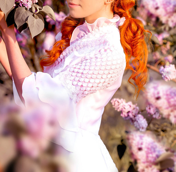 young beautiful red-haired Caucasian girl in a white dress enjoys lilac blossoms in spring park - Фото, изображение