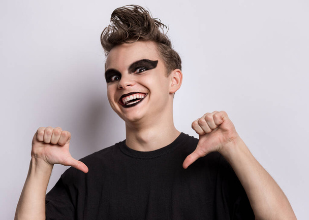 Portrait of happy teen boy with spooking make-up pointing thumbs on himself, on grey background. Smiling teenager in style of punk goth dressed in black t-shirt points fingers at oneself. - Zdjęcie, obraz