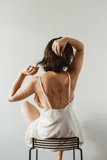 Young beautiful woman in silk underwear sitting on black stool on white background. Sexy fashion concept. - Φωτογραφία, εικόνα