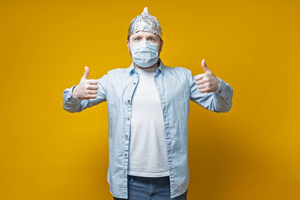 Man protected himself from the virus, put on a medical mask and a tin foil hat, he makes a thumbs up gesture and looks joyfully. - Fotoğraf, Görsel