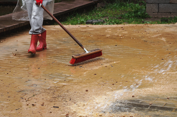 Mujer (solo piernas visibles) en botas de goma rojas limpiando pavimento de piedra cubierto de arena y agua con cepillo de cerdas rojas ásperas
 - Foto, imagen