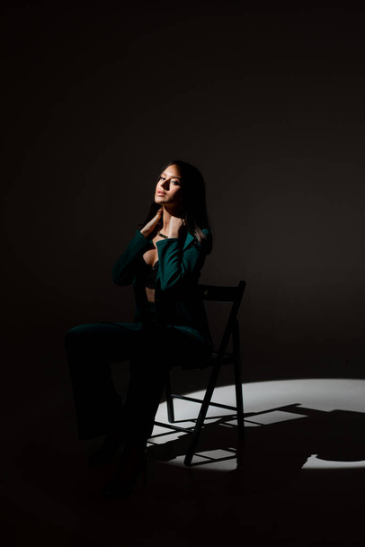Alacakaranlıkta siyah sandalyede oturan güzel esmer kadının stüdyo fotoğrafı. Yüzüne bir ışık huzmesi vuruyor.. - Fotoğraf, Görsel
