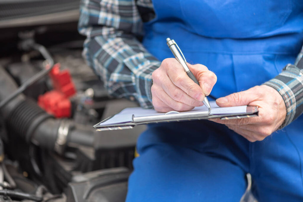 Araba tamircisi bir araba motorunu kontrol ediyor ve panoya yazı yazıyor. - Fotoğraf, Görsel