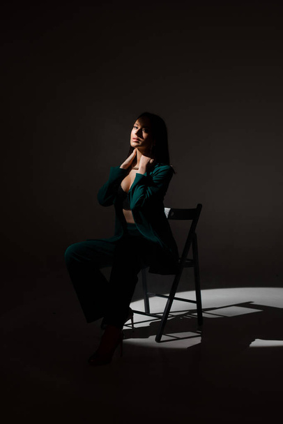 Studiová fotka krásné brunetky v soumraku sedící na černém křesle. Paprsek světla zasáhne její tvář. - Fotografie, Obrázek