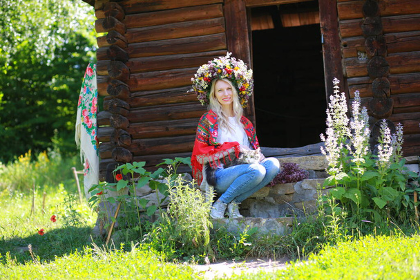 Verandada oturan genç ve güzel bir kız. Tahta bir evin yanında. Taze çiçeklerden bir çelenk ve omuzlarında kırmızı bir eşarp, Ukrayna ulusal etnik tarzı. - Fotoğraf, Görsel