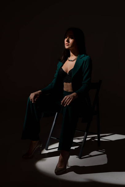 Studiofoto einer hübschen brünetten Frau in der Dämmerung, die auf einem schwarzen Stuhl sitzt. Ein Lichtstrahl trifft ihr Gesicht. - Foto, Bild