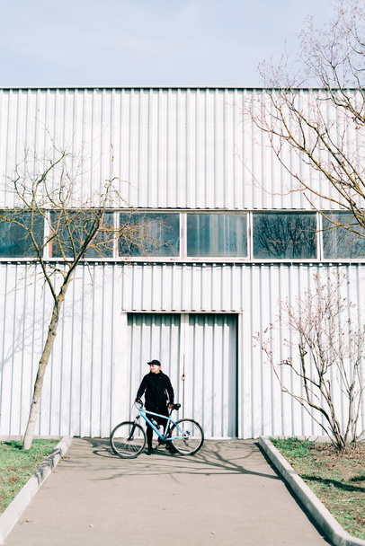 a young man in black sportswear holds a Bicycle standing next to him, against the background of a light hangar - Fotoğraf, Görsel
