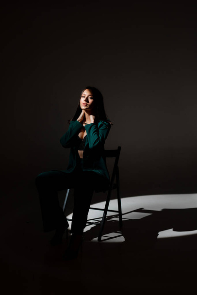Studio zdjęcie ładnej brunetki o zmierzchu siedzącej na czarnym krześle. Promień światła uderza w jej twarz.. - Zdjęcie, obraz