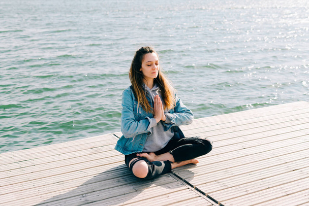 ein junges schönes Mädchen sitzt auf einem Steg in der Nähe des Sees und praktiziert Meditation - Foto, Bild