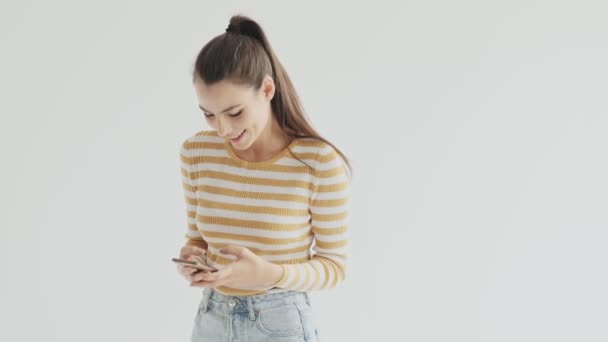 A happy laughing young woman is using her phone standing over white background in studio - Metraje, vídeo