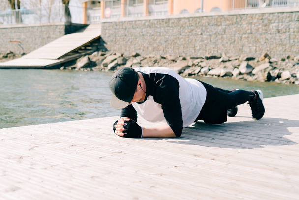 湖の近くの桟橋でスポーツ黒の服を着た若い男が春の日に板でスポーツ演習をしている - 写真・画像