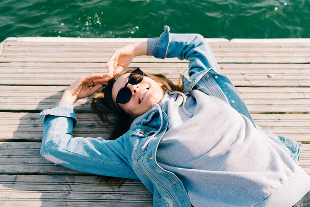 a young pretty girl in a denim jacket and glasses is lying on a pier near the lake - Fotoğraf, Görsel