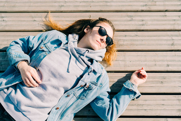 uma jovem menina bonita em uma jaqueta de ganga e óculos está deitado em um cais perto do lago
 - Foto, Imagem