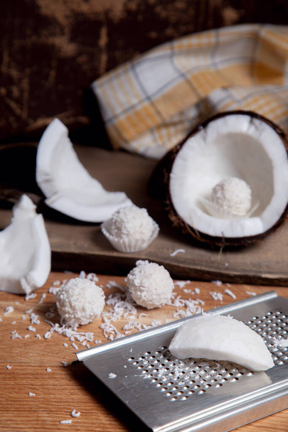 Close up of coconut with white pulp, coconut chip and sweet chocolate coconut truffles. Coconut shell, coconut flakes, small pieces of crashed nut, grater with coconut chip and white chocolate coconut sweets on a wooden background. - Foto, Imagem