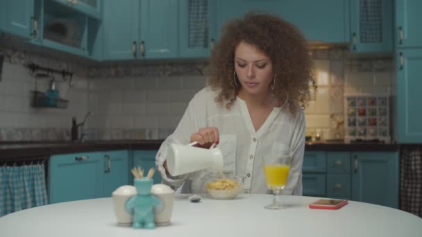 Young 20s curly hair woman eating breakfast alone on the kitchen. Female pouring vegan milk into corn flakes sitting at dining table at home.  - Filmati, video