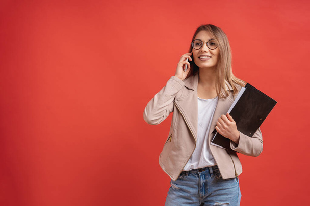 Young smiling student or intern in eyeglasses, talking on the phone, standing with a folder in hands over red background in the studio. - Foto, Imagen
