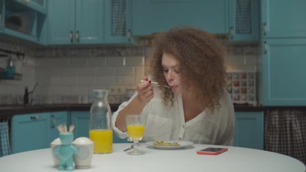 Young 20s curly hair woman eating meal alone on the kitchen. Female eating dinner and drinking orange juice at home.  - Felvétel, videó