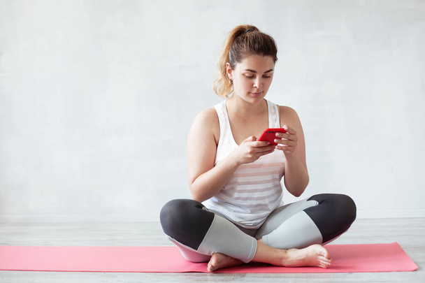 Lovely sporty woman sitting on a yoga mat with smartphone - Foto, Imagen