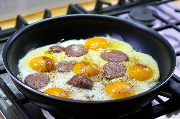 Жареные яйца с колбасой и приправами в черной сковороде на газовой плите. Вкусный и питательный завтрак для семьи. Выборочный фокус. Вид крупным планом
 - Фото, изображение