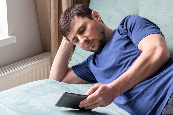 Odpoczywający mężczyzna w niebieskiej koszulce leży na zielonej sofie przy oknie z książką elektroniczną w jednej ręce, a w drugiej pod głową. Brodaty mężczyzna czyta e-booka - Zdjęcie, obraz