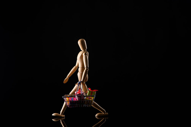 Bambola di legno che imita camminare con regali nel carrello della spesa su sfondo nero
 - Foto, immagini