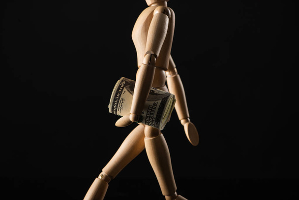 Ξύλινη κούκλα που μιμείται το περπάτημα με χαρτονομίσματα δολαρίου απομονωμένη σε μαύρο  - Φωτογραφία, εικόνα