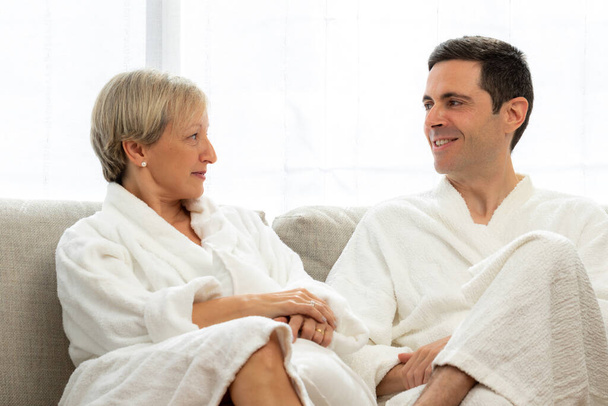 Close up portrait of middle aged couple in white bathrobes at home. Couple looking at each other on couch. - 写真・画像