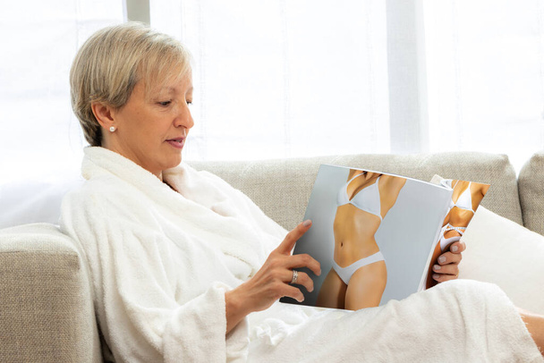 Side view portrait of middle aged woman in white bathrobe reading on couch. Woman looking at healthcare treatment magazine - Foto, Bild