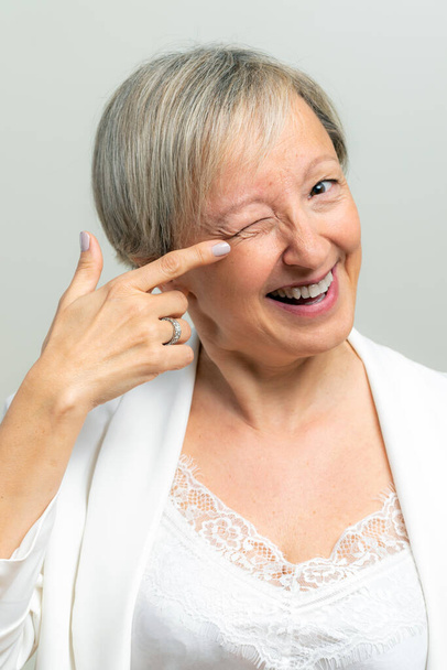 Close up studio portrait of smiling middle age woman pointing with finger at wrinkles next to eye. Woman indicating at face with one eye closed. - Photo, Image