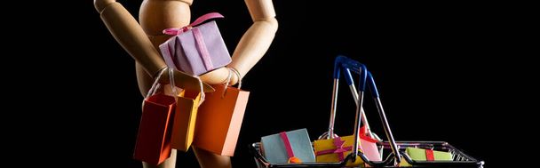 Holzpuppe mit Einkaufstaschen und Geschenken im Warenkorb isoliert auf schwarzer, panoramischer Aufnahme - Foto, Bild