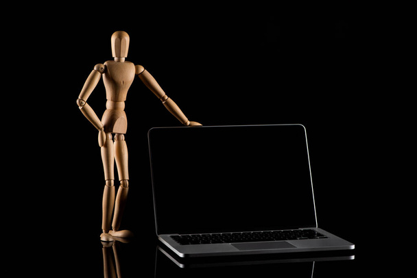 Muñeca de madera en posición de pie con la mano en la cadera cerca del ordenador portátil en negro
  - Foto, imagen