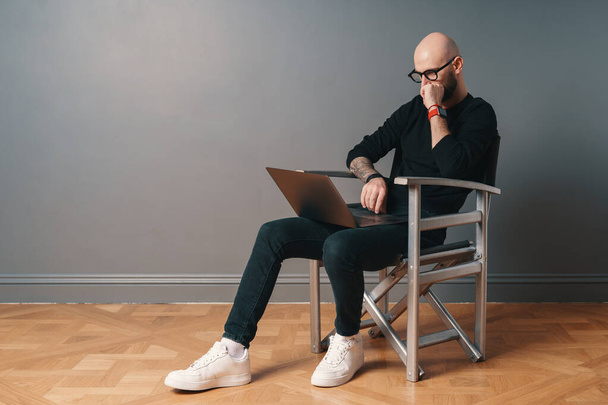 Moderni valkoihoinen mies, jolla on parta ja lasit rentouttava studio sisustus, istuu hopea tuoli, harmaa tausta ja surffaus internet kannettavan tietokoneen tietokoneella. Guy nauttia online viestintä käyttäen gadget
. - Valokuva, kuva