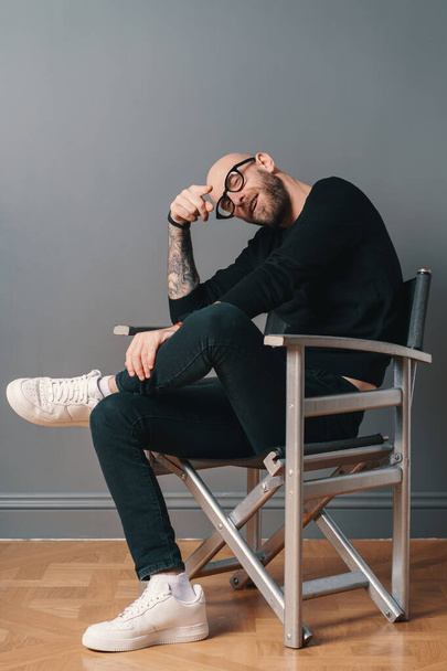 A modern, stylish man sitting leg over leg, in a confident pose on a silver chair in the studio on a gray wall background. He wears a beard, glasses, black casual clothes and white sneakers. - 写真・画像