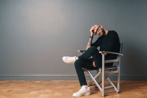 A modern, stylish man sitting leg over leg, in a confident pose on a silver chair in the studio on a gray wall background. He wears a beard, glasses, black casual clothes and white sneakers. - Foto, Bild