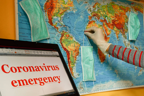 ラップトップ上の世界地図や単語の緊急コロナウイルスに医療マスクをぶら下げながらラテックス手袋で手 - 写真・画像