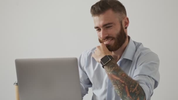 A confident young bearded man wearing an office style is using his laptop computer sitting at the table over white wall - Πλάνα, βίντεο