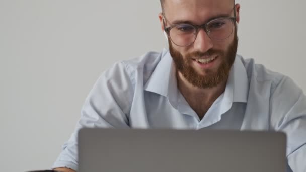 A positive smiling bearded young man in eyeglasses and wireless earphones is talking to someone in a video chat while using a laptop computer over white wall - Záběry, video