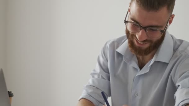 A confident bearded young man wearing wireless earphones and glasses is working with the documents while sitting at the table isolated over white wall - Video