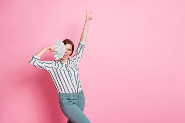 Yeah i millionaire. Delighted positive girl cool she win lottery casino dollars cover face money fan scream show v-sign wear white striped shirt pants trousers isolated pink color background - Foto, Bild