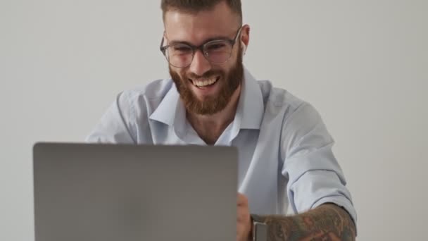 A happy smiling young man wearing wireless earphones and eyeglasses is having a video chat using his laptop computer isolated over white wall - Felvétel, videó
