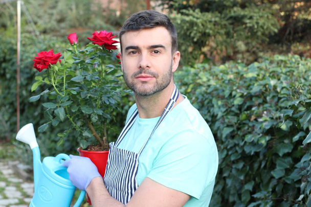 γκρο πλαν πορτρέτο του όμορφου νέου άνδρα με κόκκινα τριαντάφυλλα και ποτιστήρι στον κήπο - Φωτογραφία, εικόνα