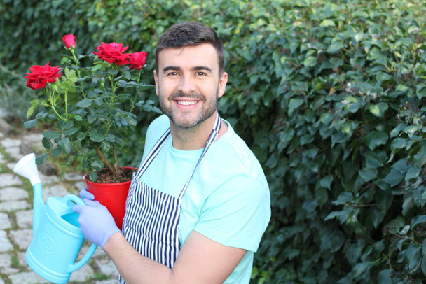 γκρο πλαν πορτρέτο του όμορφου νέου άνδρα με κόκκινα τριαντάφυλλα και ποτιστήρι στον κήπο - Φωτογραφία, εικόνα